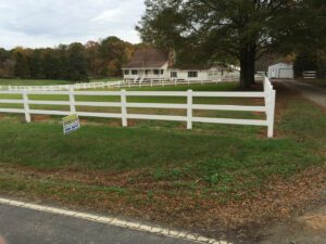 White fence along a grassy roadside with a house and trees in the background.