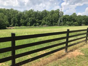 A wooden fence borders a grassy field with trees under a blue sky.