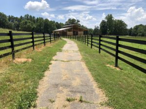 A gravel driveway leading to a barn surrounded by green fields and black fences.