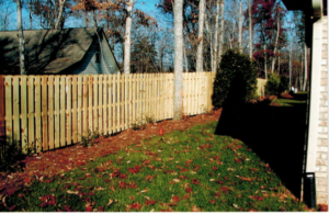 A wooden picket fence with trees and fallen leaves in the yard.