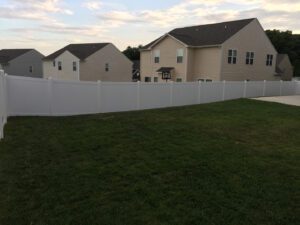 Suburban homes with a white vinyl fence enclosing a grassy yard at dusk.